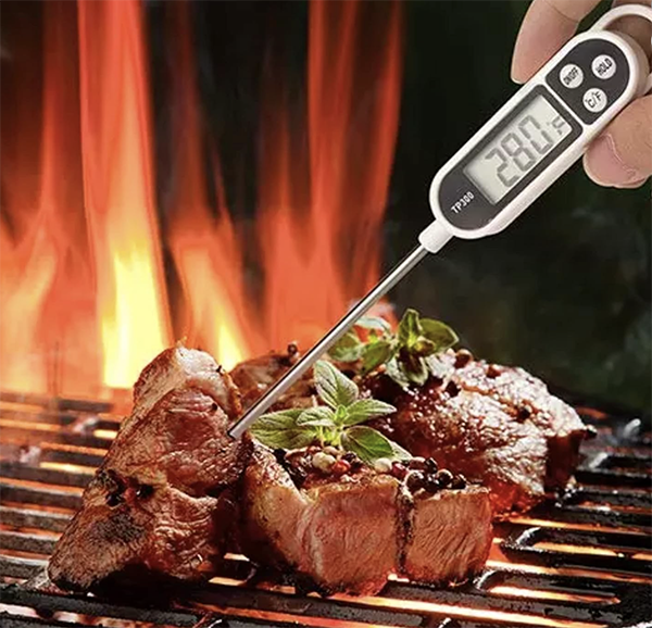 Digital thermometer inserted into a piece of meat on a grill with flames in the background